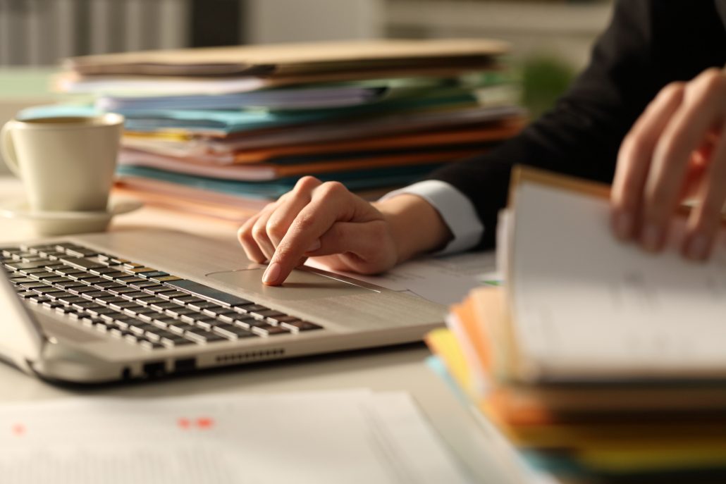 Close up of executive hands working hard late hours using laptop in the night at office checking a pile of documents
